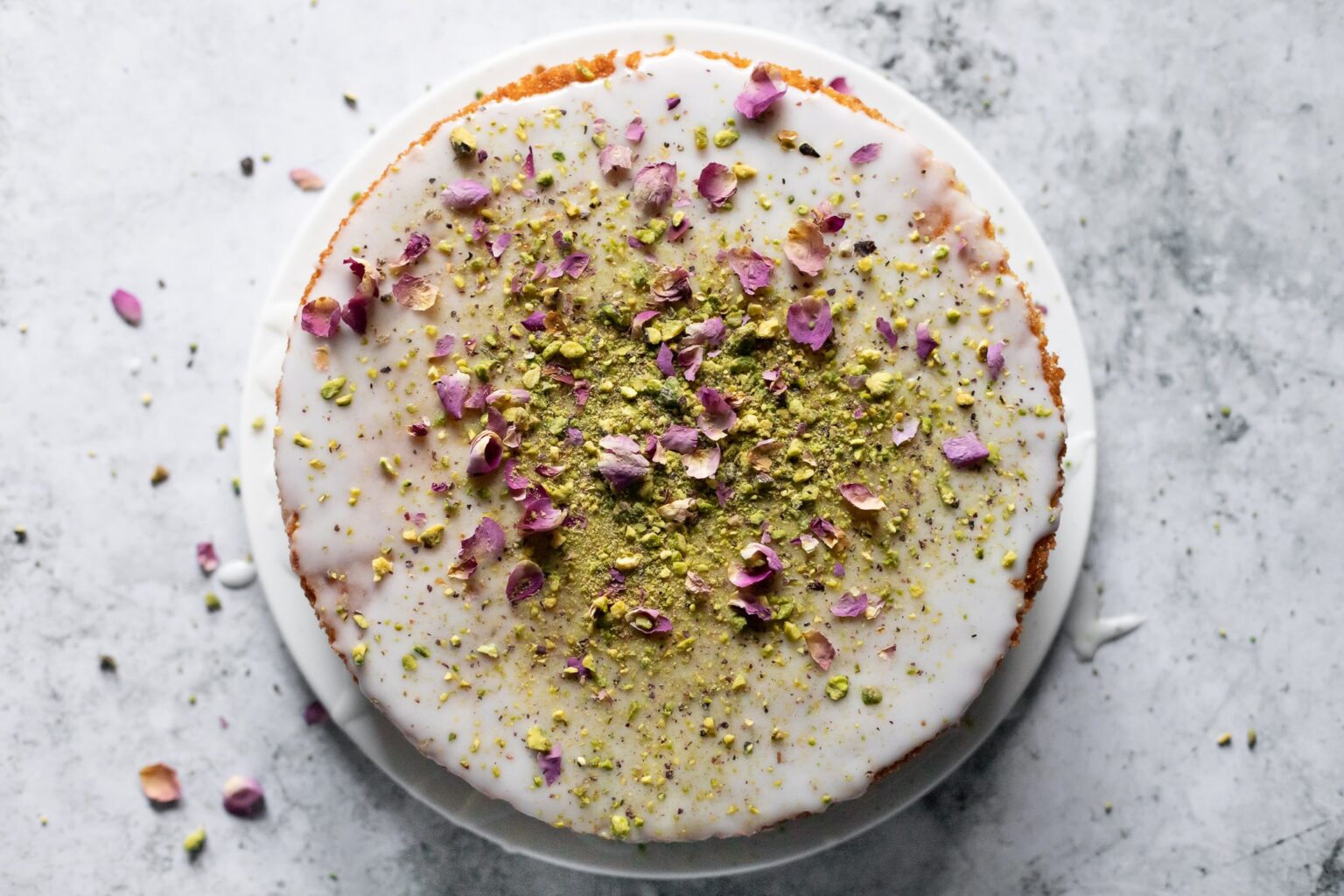 overhead photo of a cake decorated with white icing, green pistachios, and pink dried rose petals